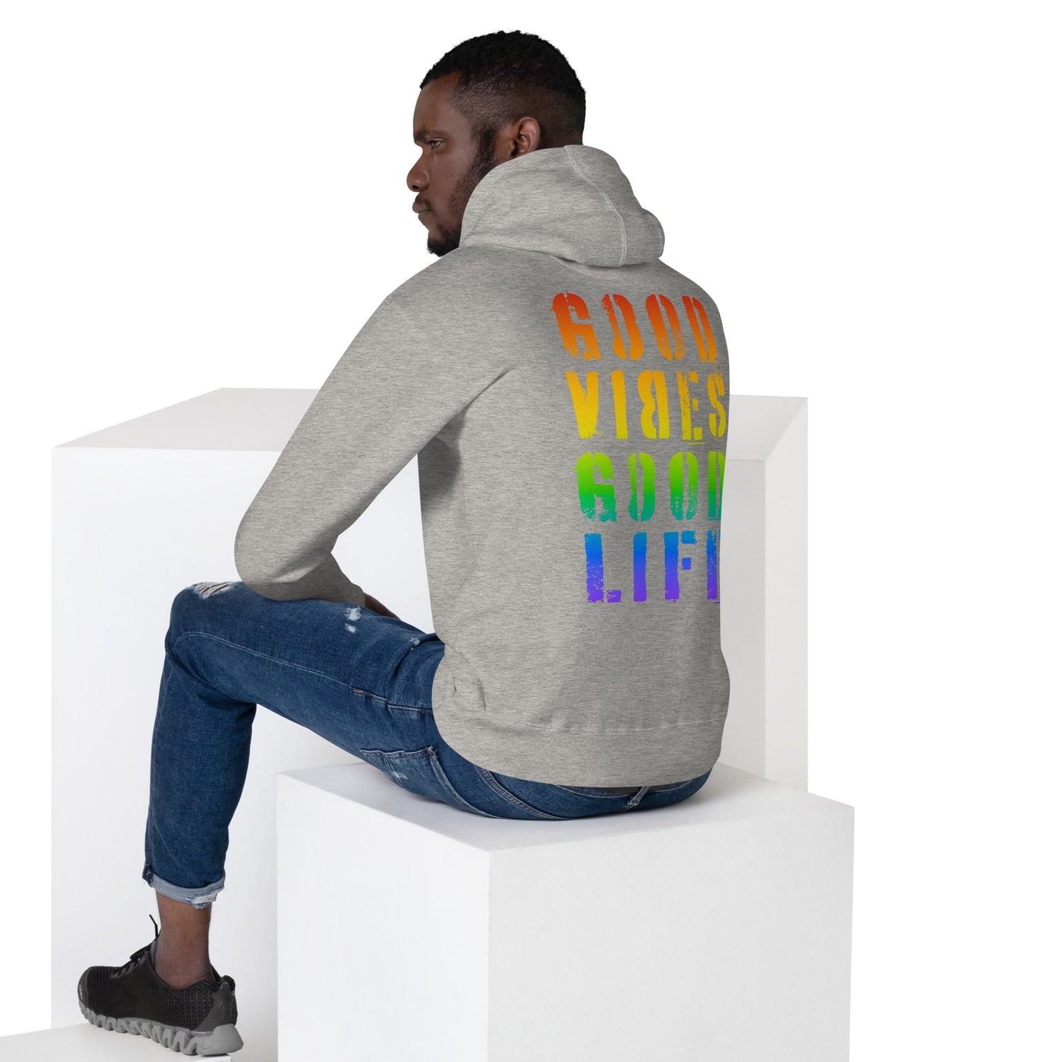 Mann in grauem Hoodie mit Regenbogenprint 'GOOD VIBES GOOD LIFE', sitzt auf weißem Würfel