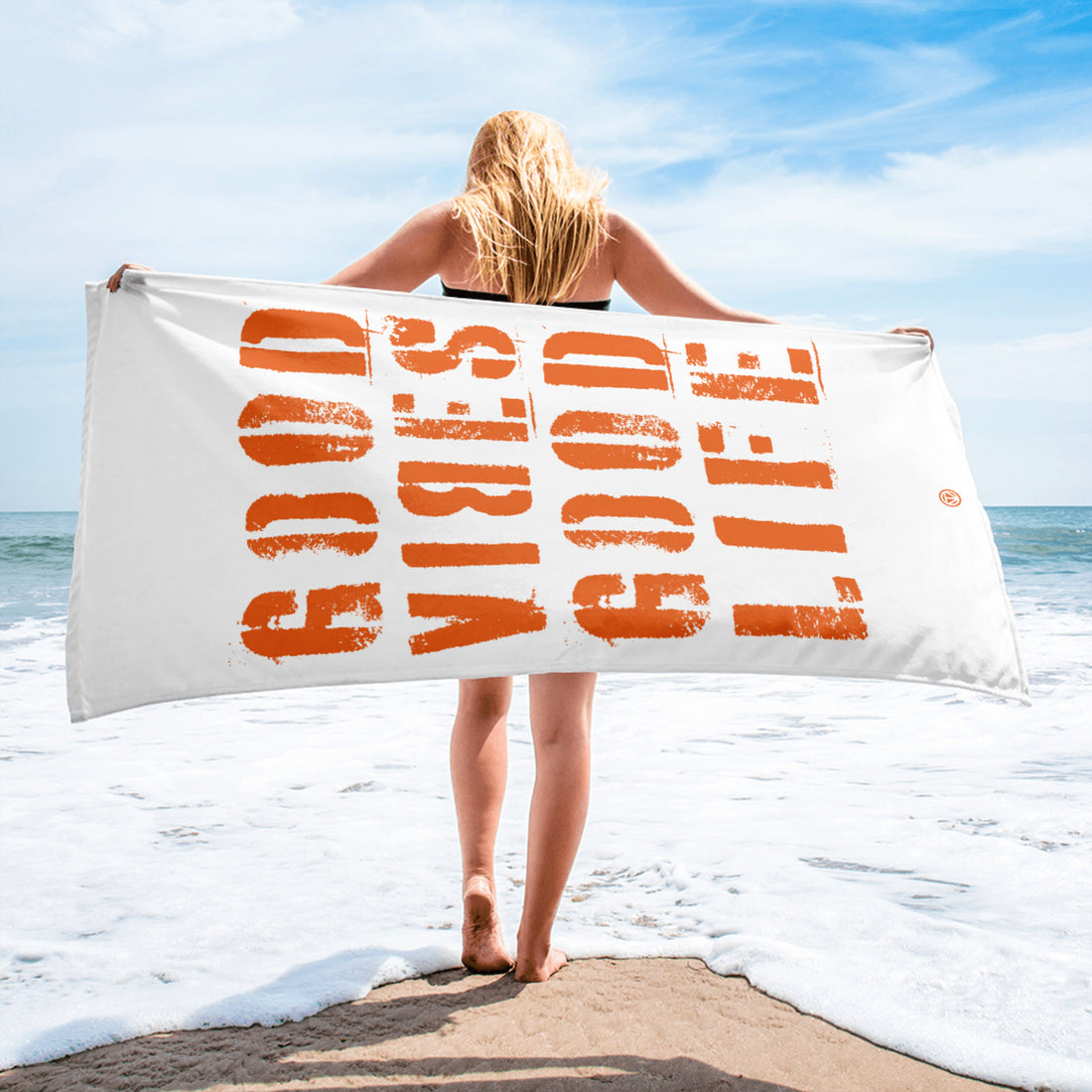 Frau am Strand hält weißes Strandtuch mit orangefarbenem Schriftzug &
