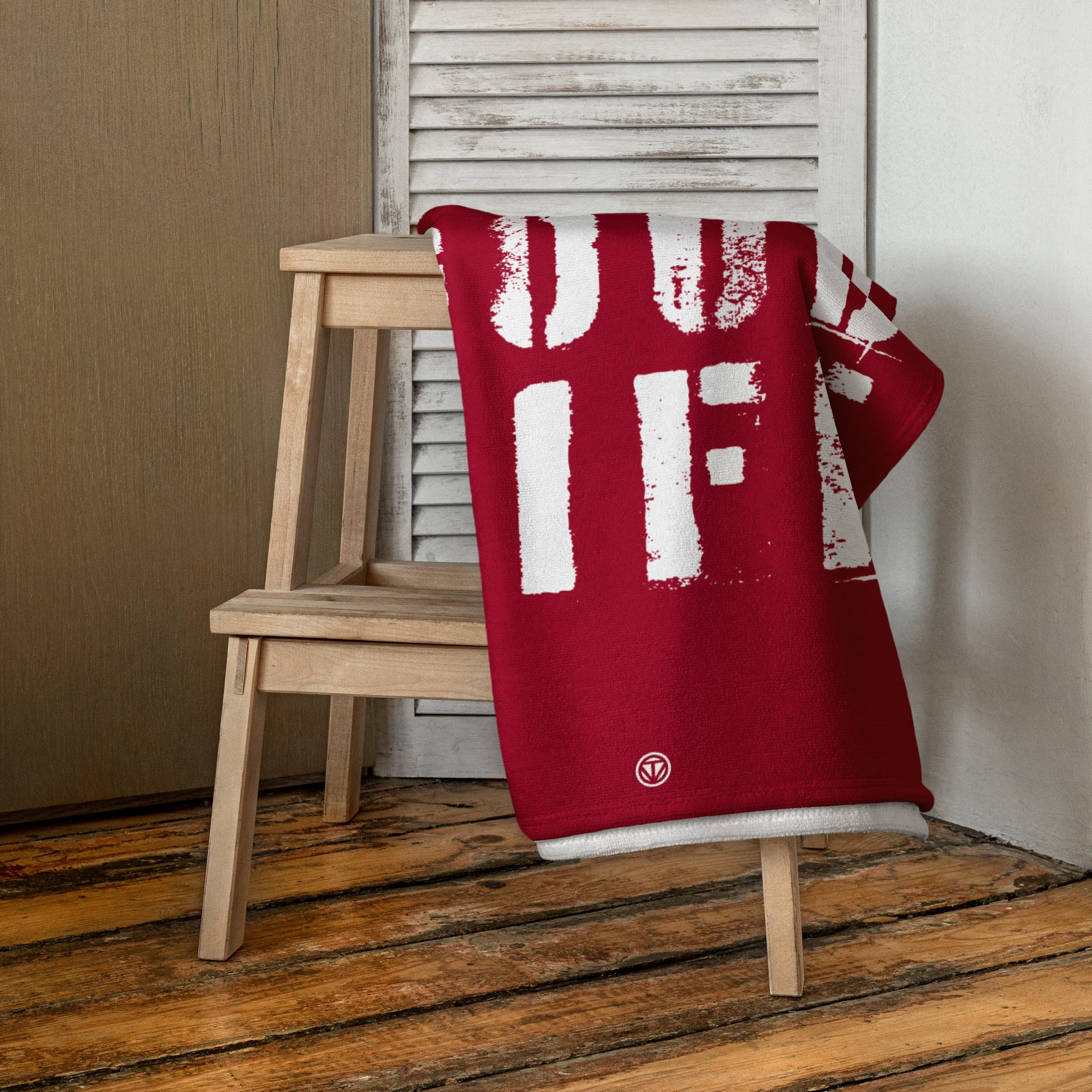 Rotes Strandtuch mit weißem Schriftmuster liegt gefaltet auf hellem Holzschemel vor weißer Lamellentür