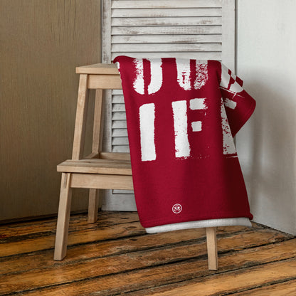 Rotes Strandtuch mit weißem Schriftmuster liegt gefaltet auf hellem Holzschemel vor weißer Lamellentür