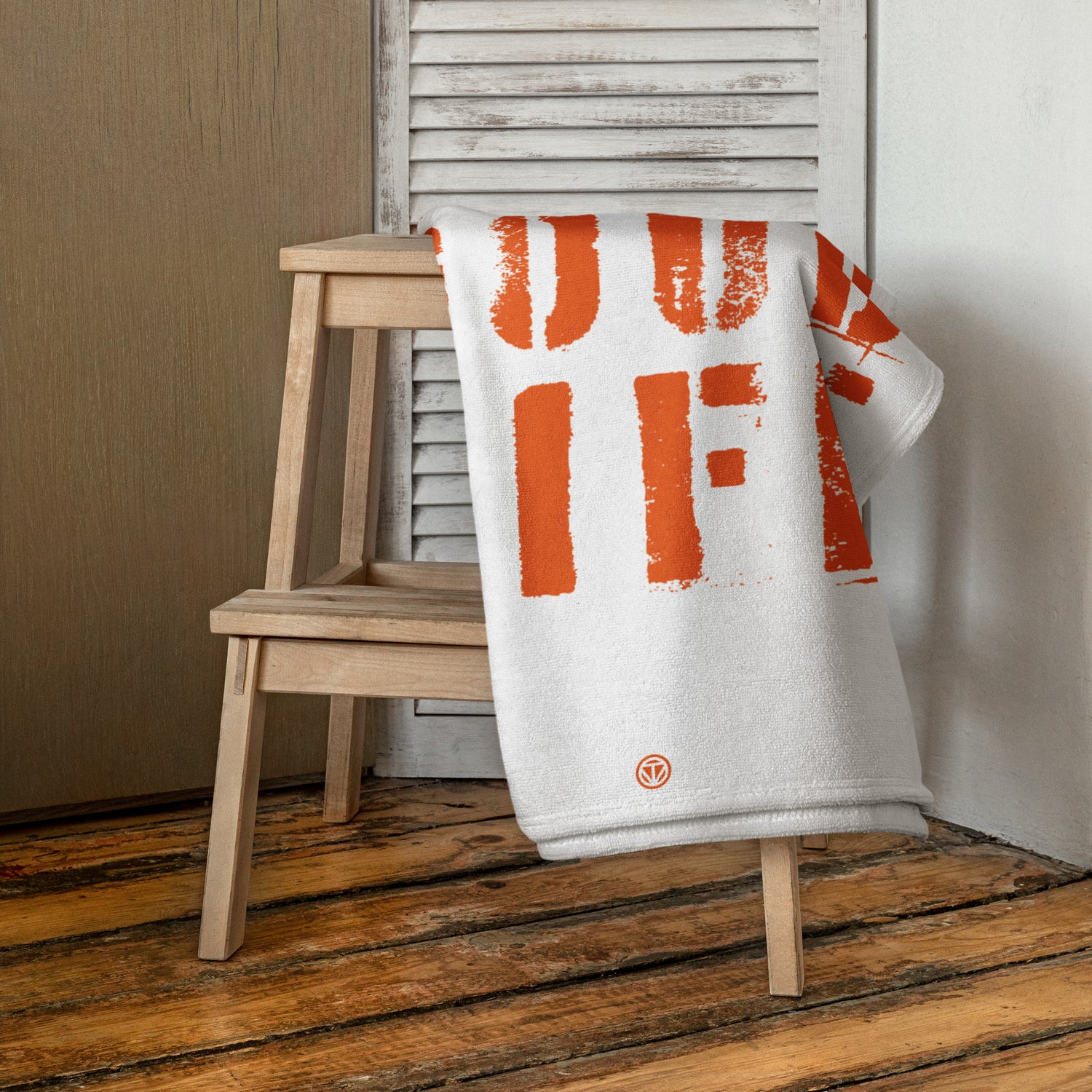 Weiße Strandtuch mit orangefarbenem Aufdruck auf Holzstuhl vor weißem Fensterladen und Holzboden